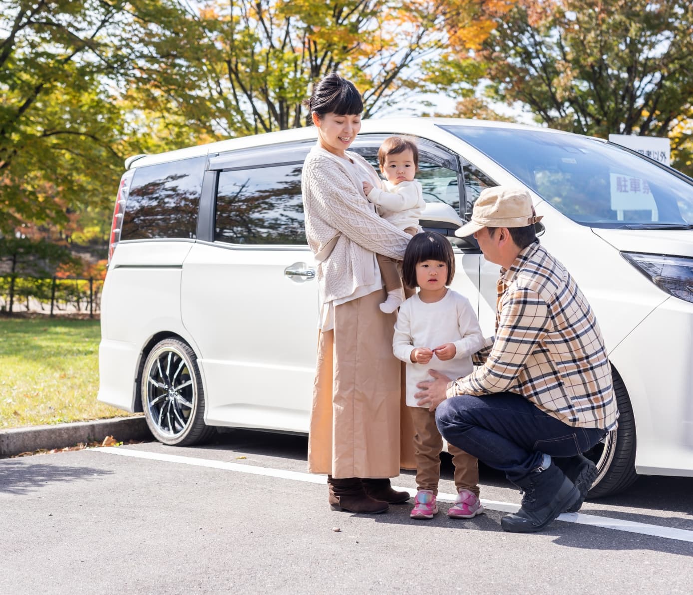 白い車の前で、幼い子供と話す若い親子の写真